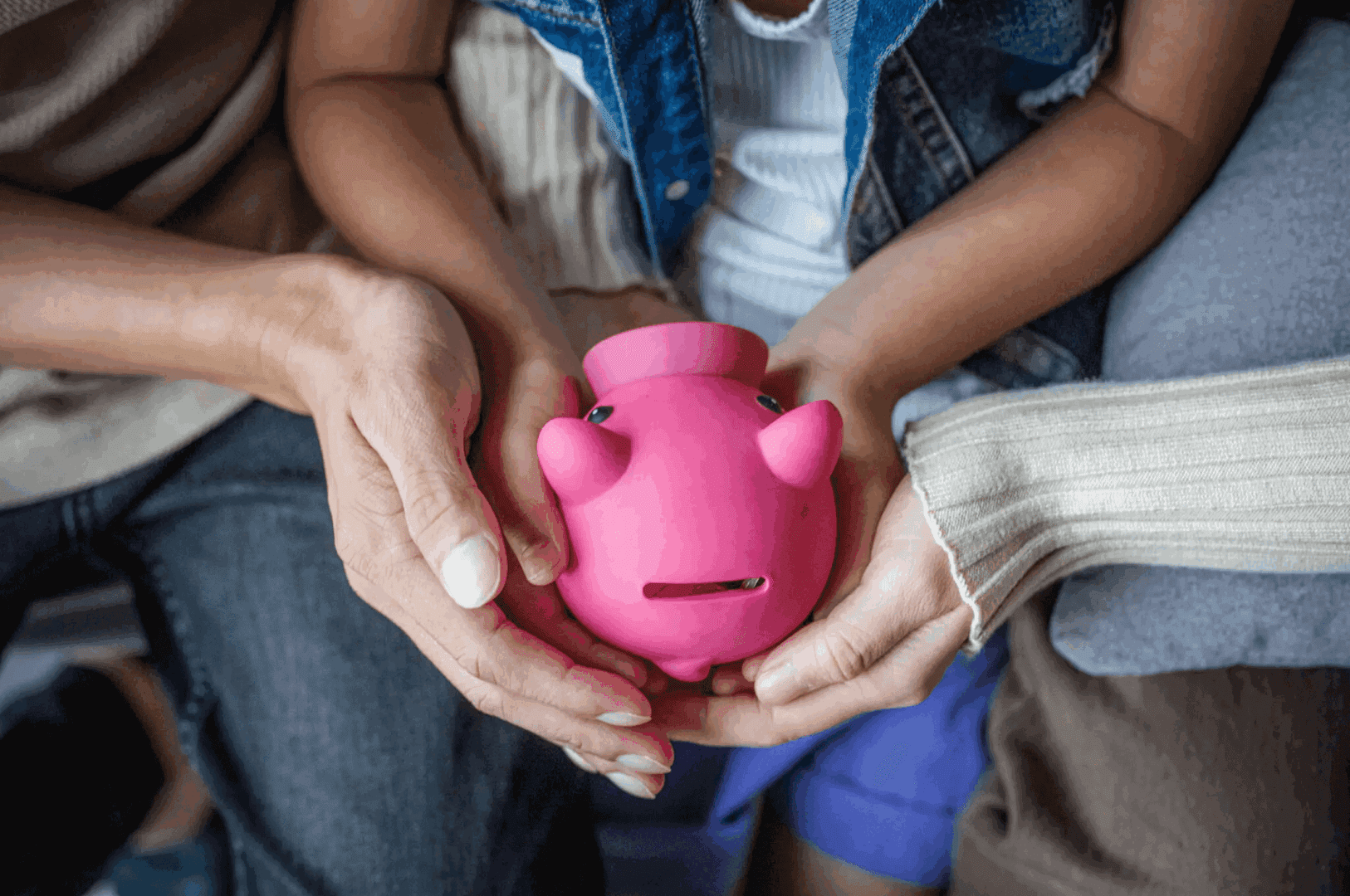 Hands of a child and adult holding a pink piggy bank together.