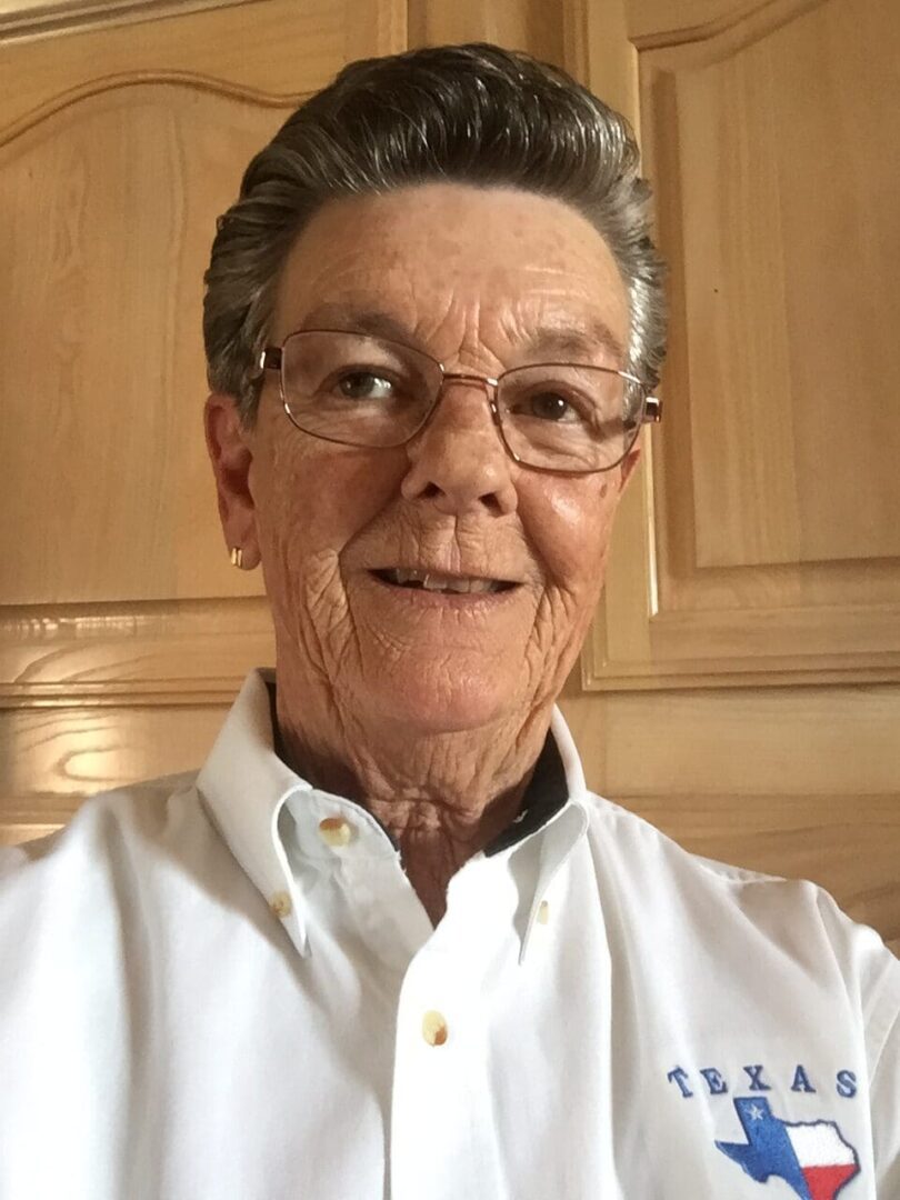 Smiling older woman wearing glasses and a white shirt in a cozy indoor setting.