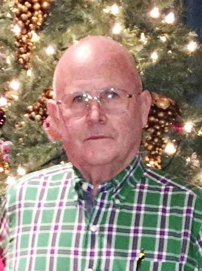 Elderly man in glasses and plaid shirt stands before a decorated Christmas tree.